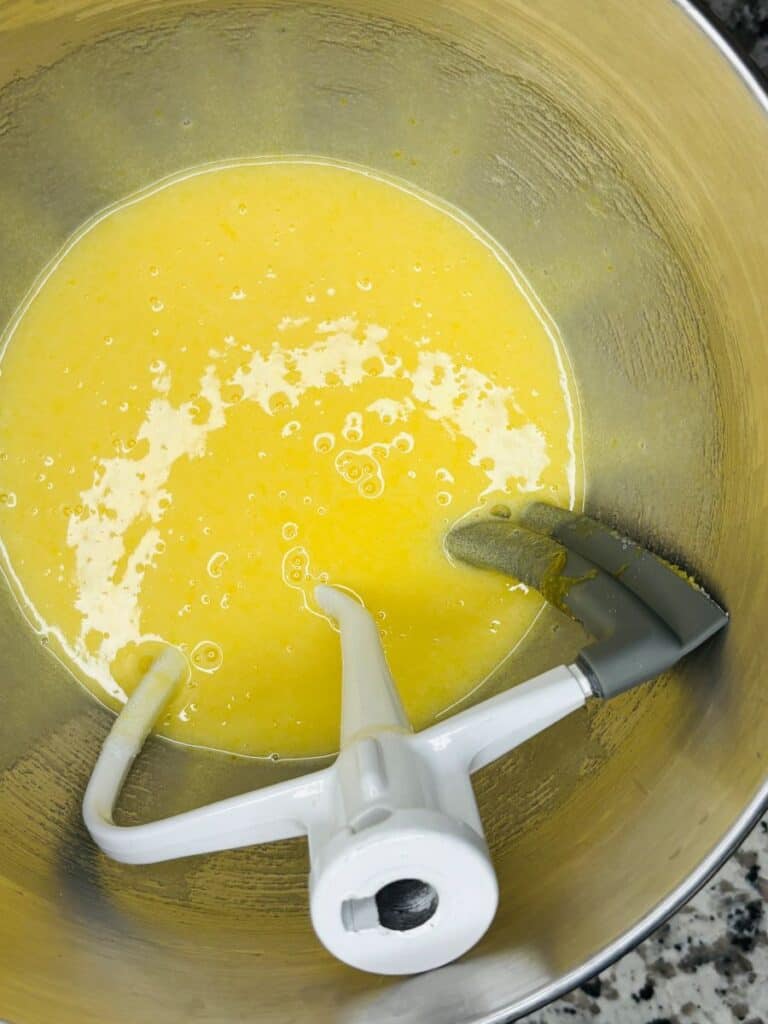 A metal mixing bowl with a stand mixer paddle attachment, containing a yellow batter being mixed. The bowl sits on a speckled countertop.