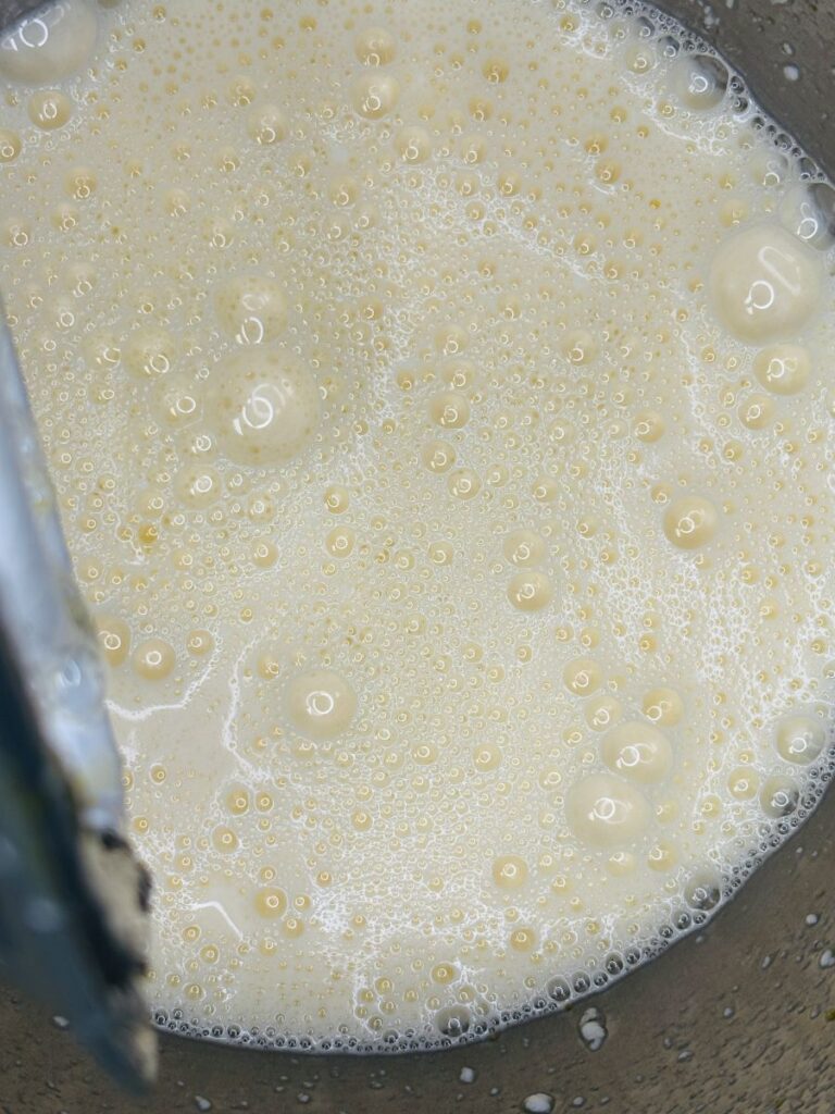 A close-up of a bowl containing a frothy, light yellow liquid mixture with bubbles on the surface, possibly batter or a beaten egg mixture.