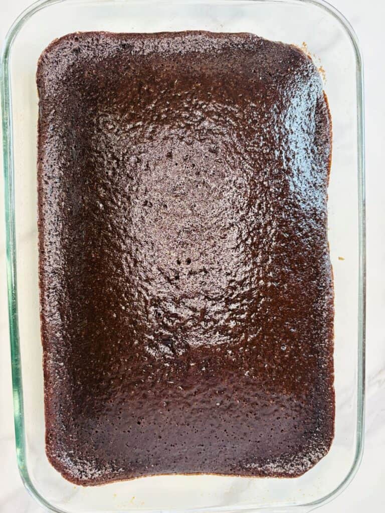 A rectangular glass baking dish filled with a freshly baked, uncut chocolate brownie, showing a shiny, slightly textured top and lightly browned edges.