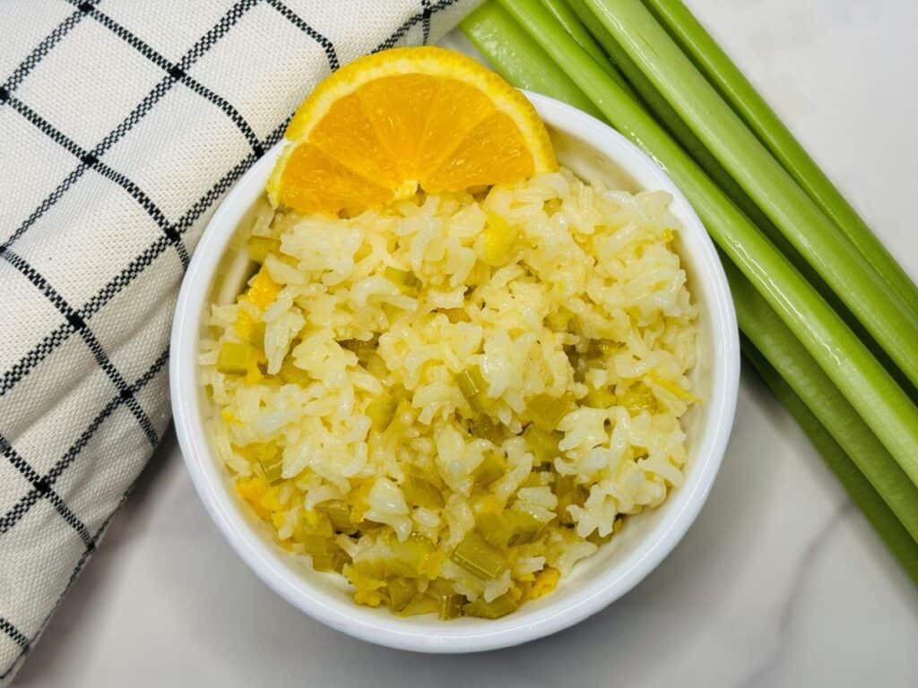 A bowl of white rice mixed with green vegetables, garnished with an orange slice, sits next to celery stalks and a checkered cloth on a white surface.