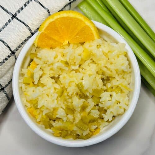 A bowl of white rice mixed with green vegetables, garnished with an orange slice, sits next to celery stalks and a checkered cloth on a white surface.