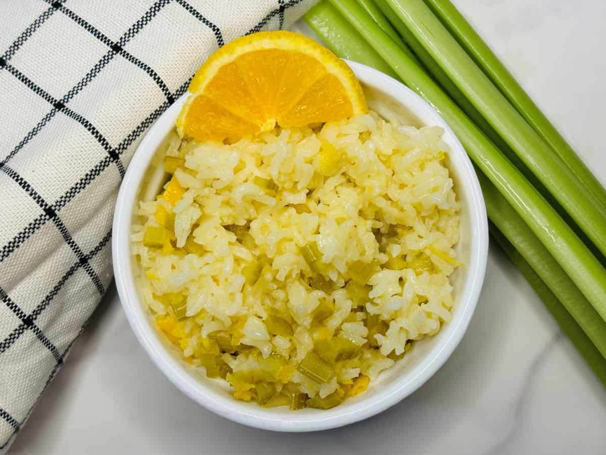 A bowl of white rice mixed with green vegetables, garnished with an orange slice, sits next to celery stalks and a checkered cloth on a white surface.