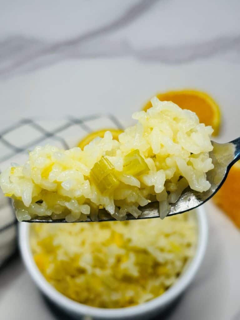 A close-up of a spoonful of cooked rice with pieces of celery, held above a bowl of the same rice dish. Sliced oranges and a checkered cloth are in the blurred background.