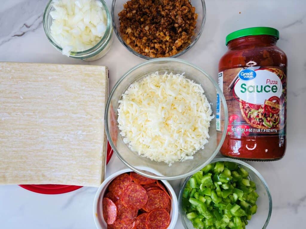 Ingredients for making pizza, including chopped onions, cooked sausage, pizza sauce, shredded cheese, green peppers, pepperoni slices, and pizza dough, all arranged in bowls and containers on a marble countertop.