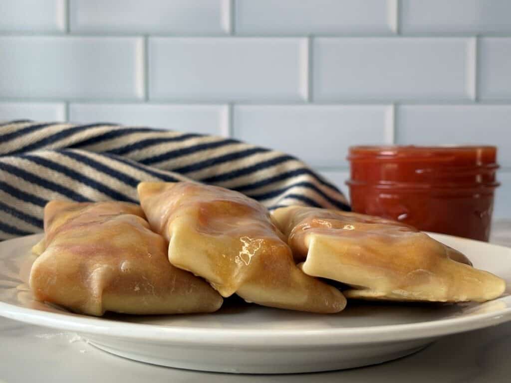 Three golden-brown pierogi on a white plate, with a jar of red sauce in the background and a striped cloth behind them, set against a white tiled wall.