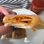 A hand holds a bitten pizza roll, showing melted cheese and sauce inside. A plate with more rolls and a jar of red sauce are in the background.