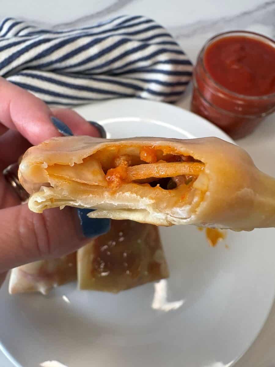 A hand holds a bitten pizza roll, showing melted cheese and sauce inside. A plate with more rolls and a jar of red sauce are in the background.