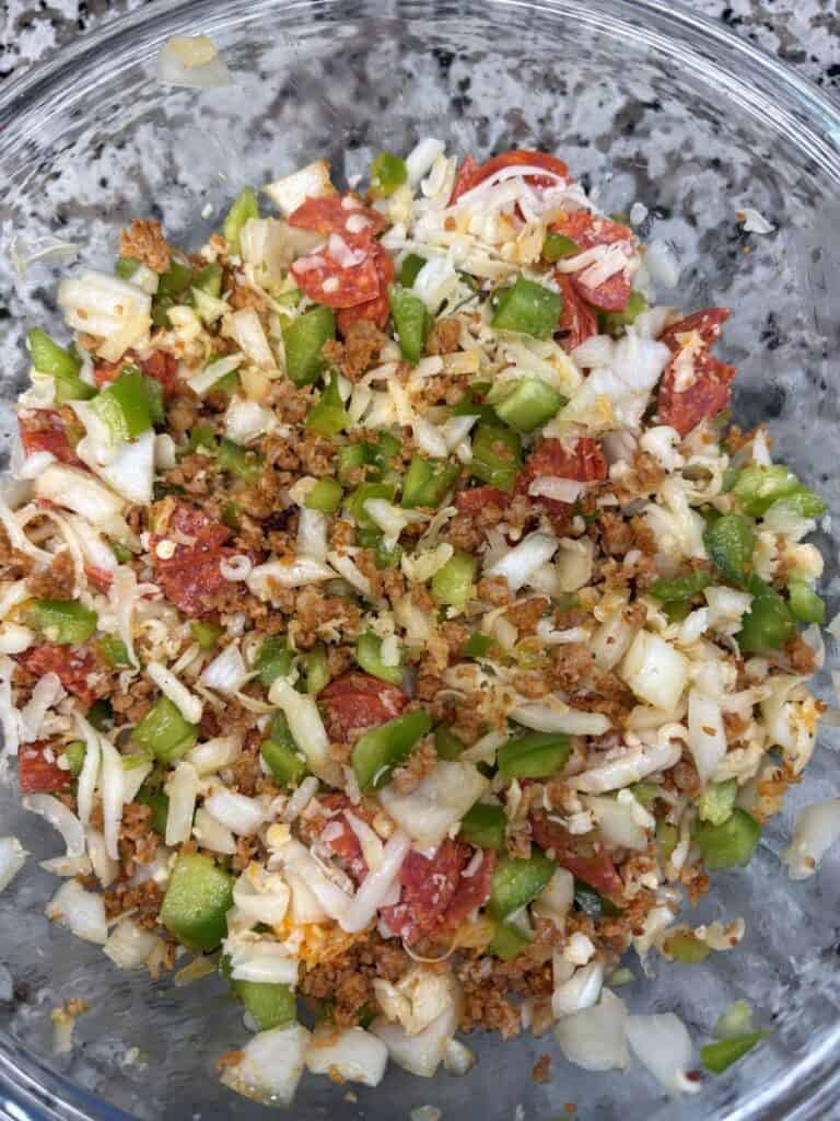 A glass bowl filled with chopped green peppers, onions, shredded cheese, diced pepperoni, and crumbled sausage on a speckled countertop.