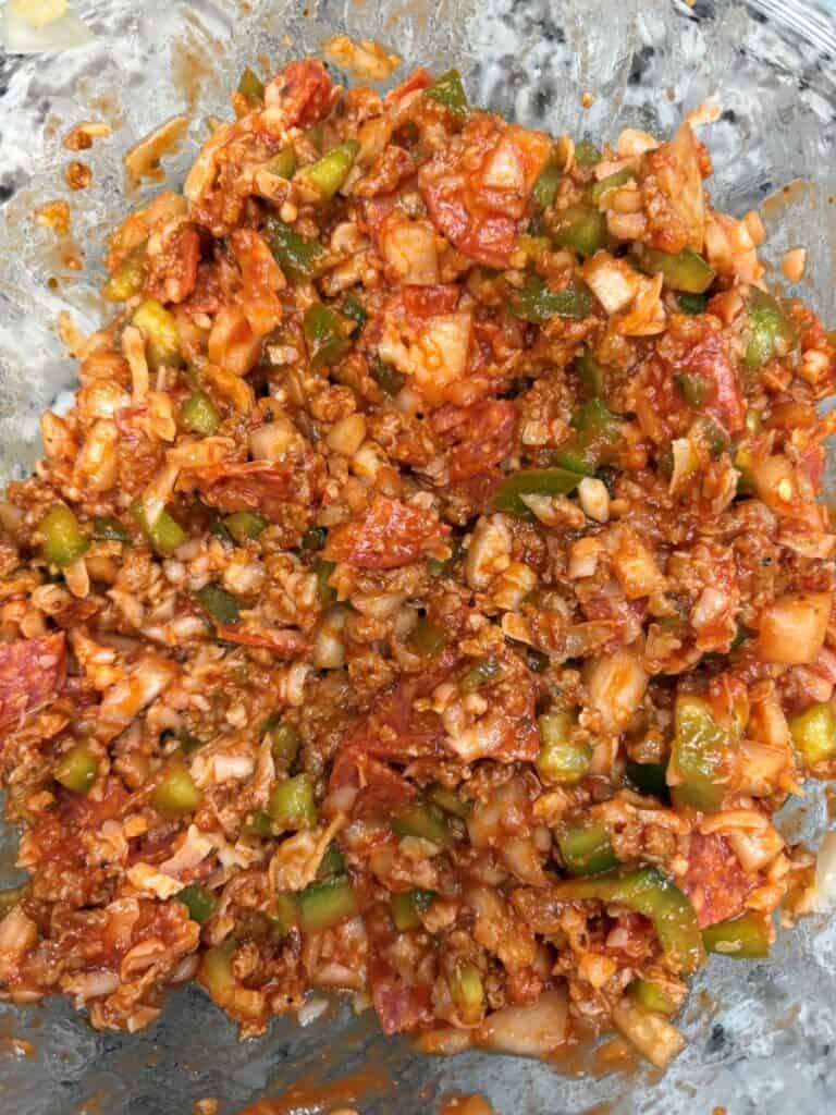 A close-up of a mixed dish containing chopped green peppers, onions, pepperoni slices, cheese, and red sauce in a glass bowl.