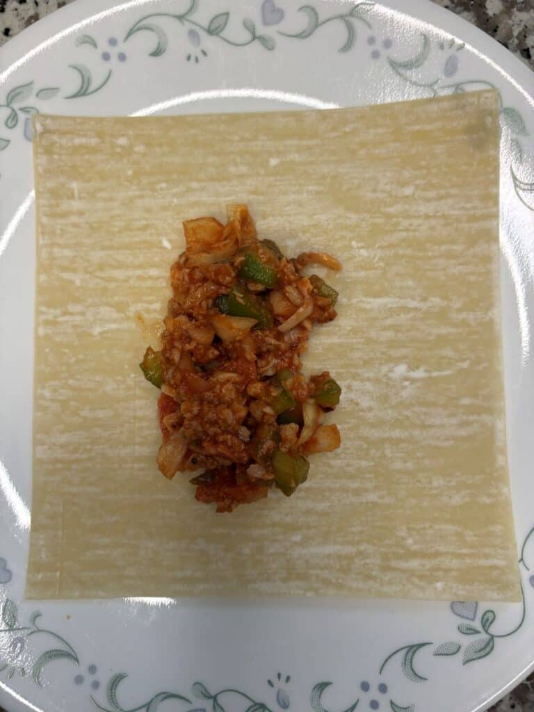 A square sheet of uncooked pasta on a white plate with a floral pattern, topped with a portion of vegetable and meat tomato sauce filling in the center.