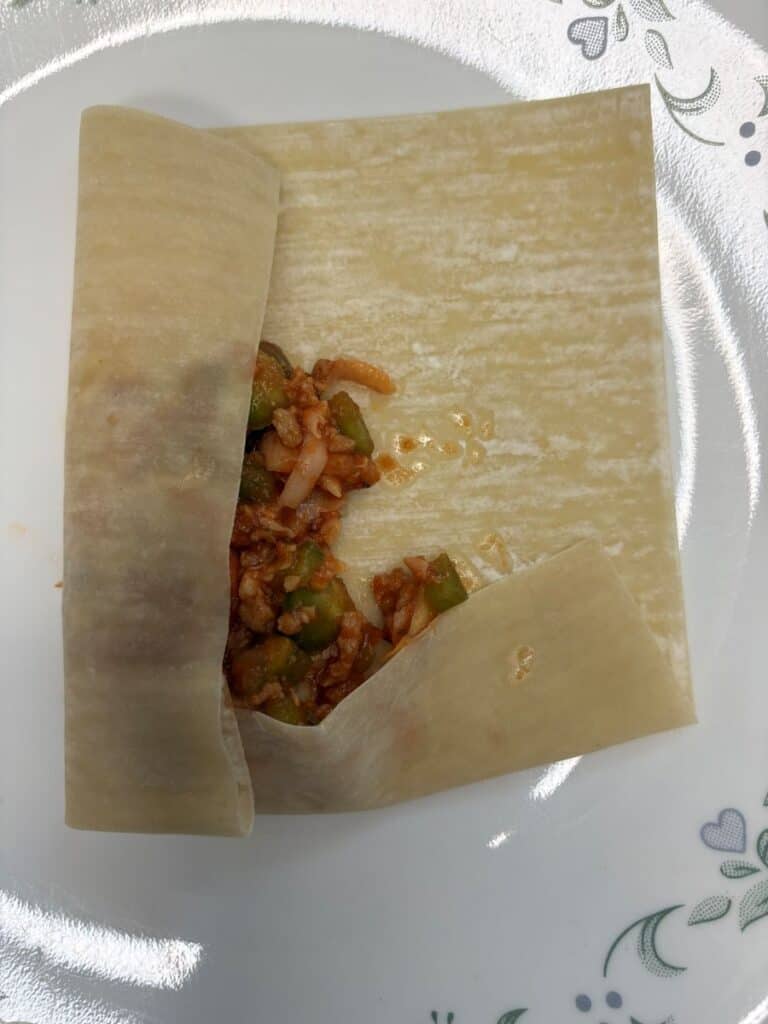 A square wonton wrapper partially folded over a filling of minced vegetables and meat on a white plate with a floral pattern.