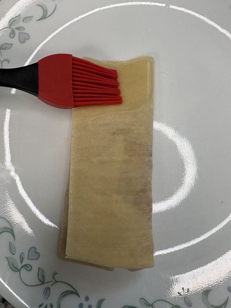 A red silicone brush is applying liquid, likely egg wash or water, to folded sheets of pastry or dough on a white plate with a floral border.