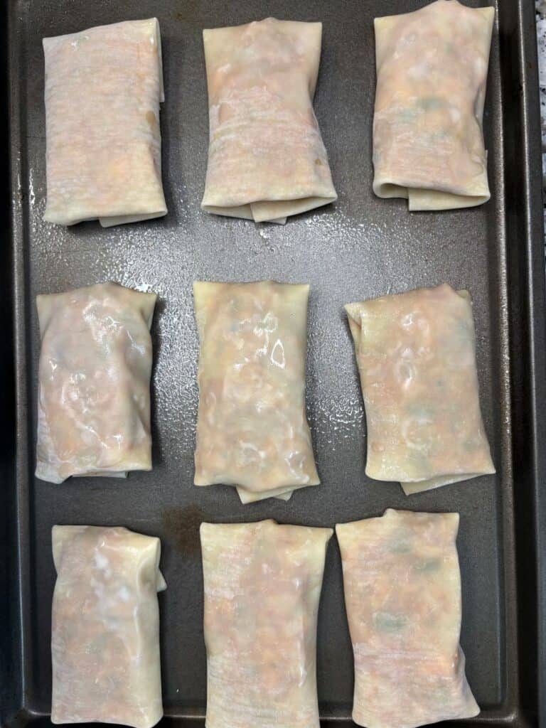 Nine uncooked spring rolls arranged in three rows on a dark baking tray, each wrapped in a translucent wrapper revealing the filling inside.