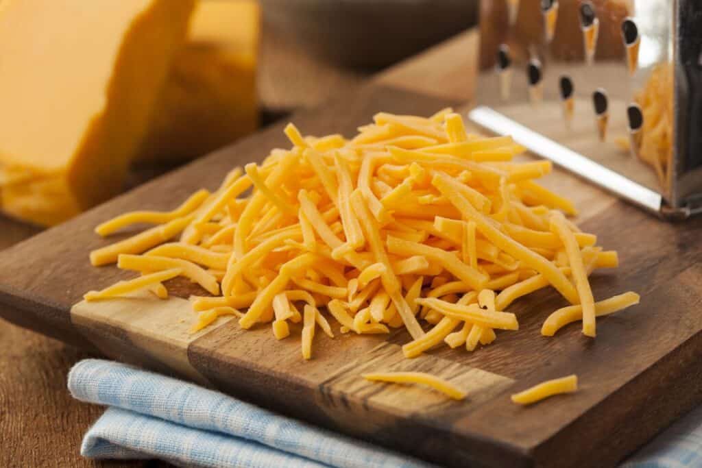 A pile of shredded cheddar cheese sits on a wooden cutting board next to a metal grater, with a block of cheese and a blue-striped cloth nearby.