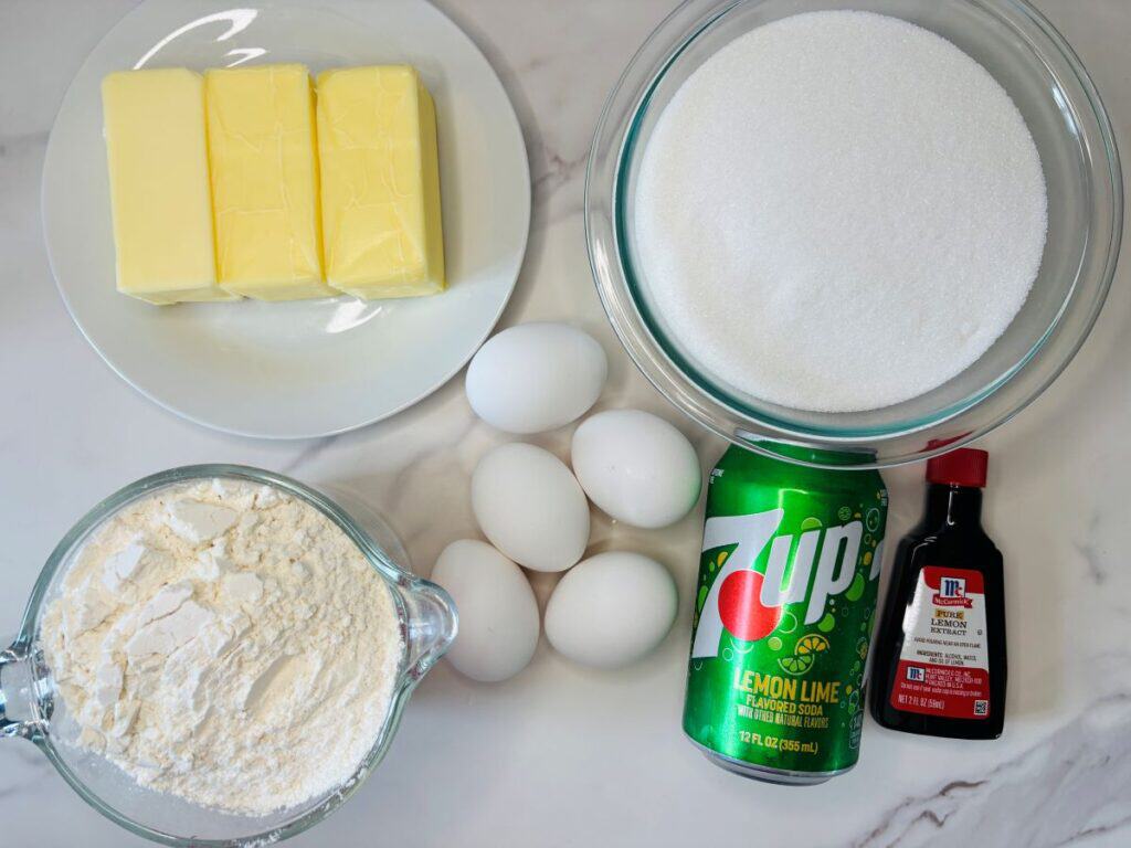 Three sticks of butter on a plate, a bowl of sugar, a bowl of flour, five eggs, a can of 7UP soda, and a bottle of vanilla extract on a white countertop.