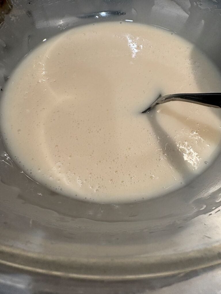 A close-up of a glass bowl filled with a creamy white liquid, possibly batter or custard, with a metal spoon partially submerged in it.