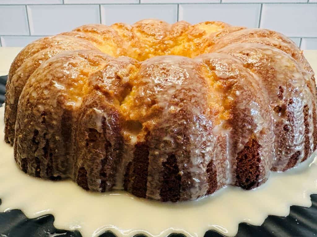 A glazed bundt cake sits on a black plate, with white icing dripping down its sides. The background shows white subway tiles.