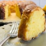 A close-up of a slice of moist lemon bundt cake with glaze on a white plate and a fork, with the rest of the cake and a lemon in the background.
