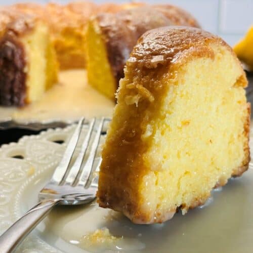 A close-up of a slice of moist lemon bundt cake with glaze on a white plate and a fork, with the rest of the cake and a lemon in the background.