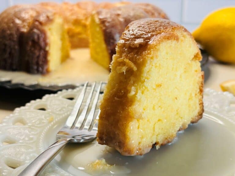 A close-up of a slice of moist lemon bundt cake with glaze on a white plate and a fork, with the rest of the cake and a lemon in the background.
