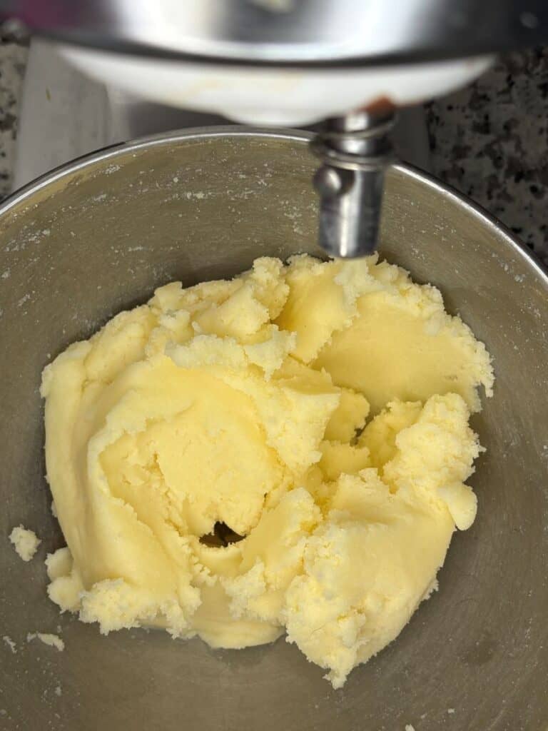 A metal mixing bowl with pale yellow cookie dough or batter being mixed in a stand mixer. The dough has a smooth, creamy texture.
