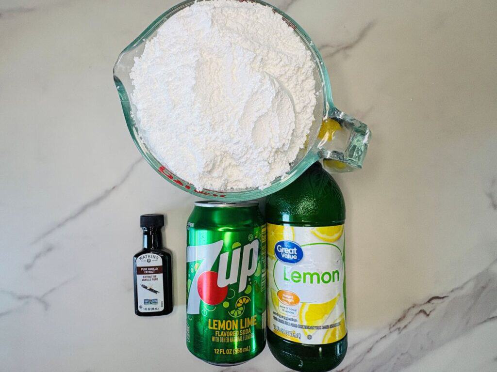 A measuring cup of flour, a small bottle of vanilla extract, a can of 7UP, and a bottle of lemon juice on a marble countertop.
