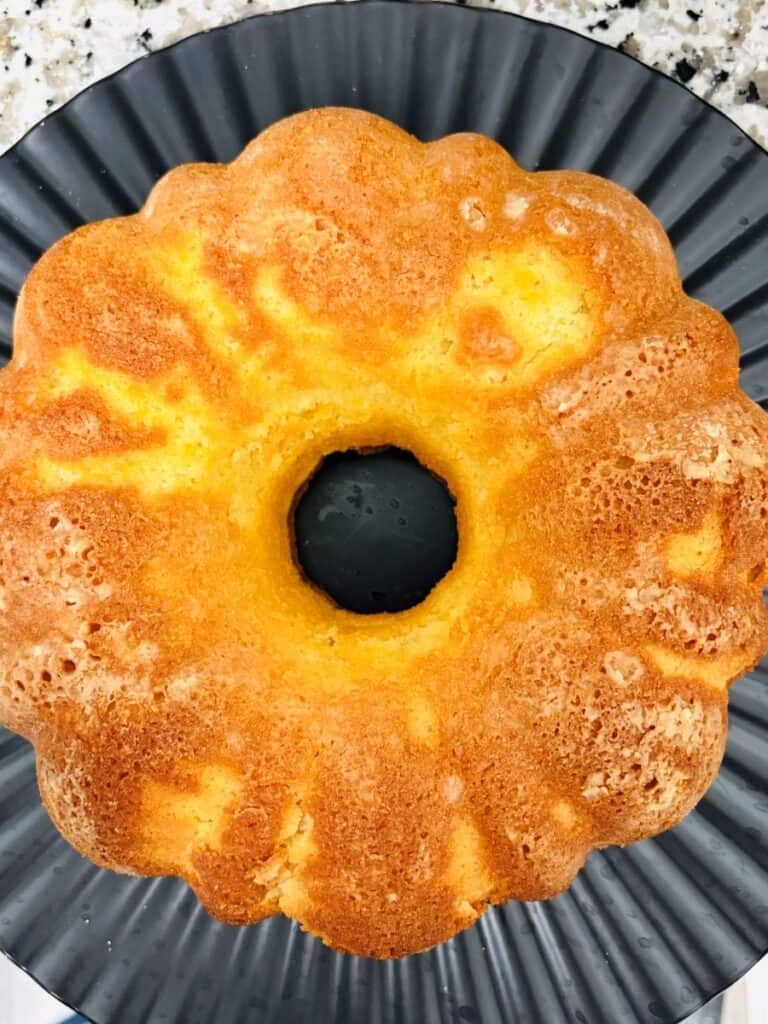 A golden-brown bundt cake sits on a dark, ridged plate, showing a light, fluffy texture with a central hole and slightly crisp edges.