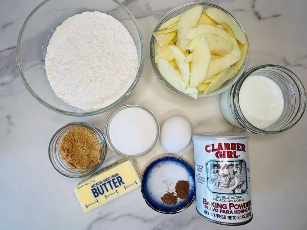 Baking ingredients on a marble surface: flour, sliced apples, milk, granulated sugar, brown sugar, an egg, butter, salt with spices, and a can of Clabber Girl baking powder.
