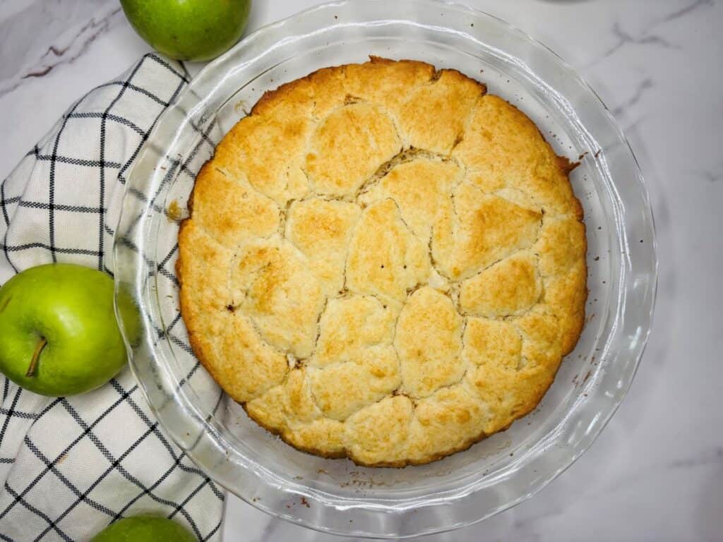 A baked apple cobbler with a golden-brown biscuit topping in a glass pie dish, surrounded by green apples and a checked kitchen towel on a marble surface.