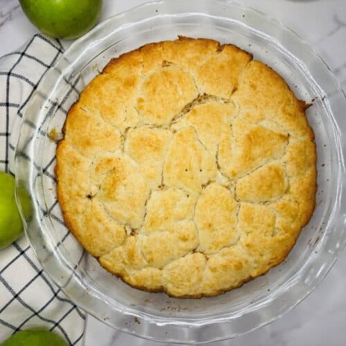 A baked apple cobbler with a golden-brown biscuit topping in a glass pie dish, surrounded by green apples and a checked kitchen towel on a marble surface.