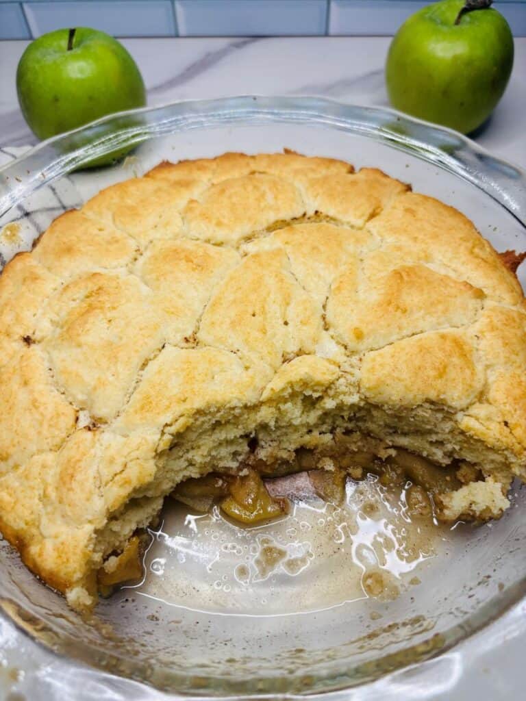 A partially eaten apple cobbler in a glass dish sits on a marble counter, with two green apples and a white tile backsplash in the background.