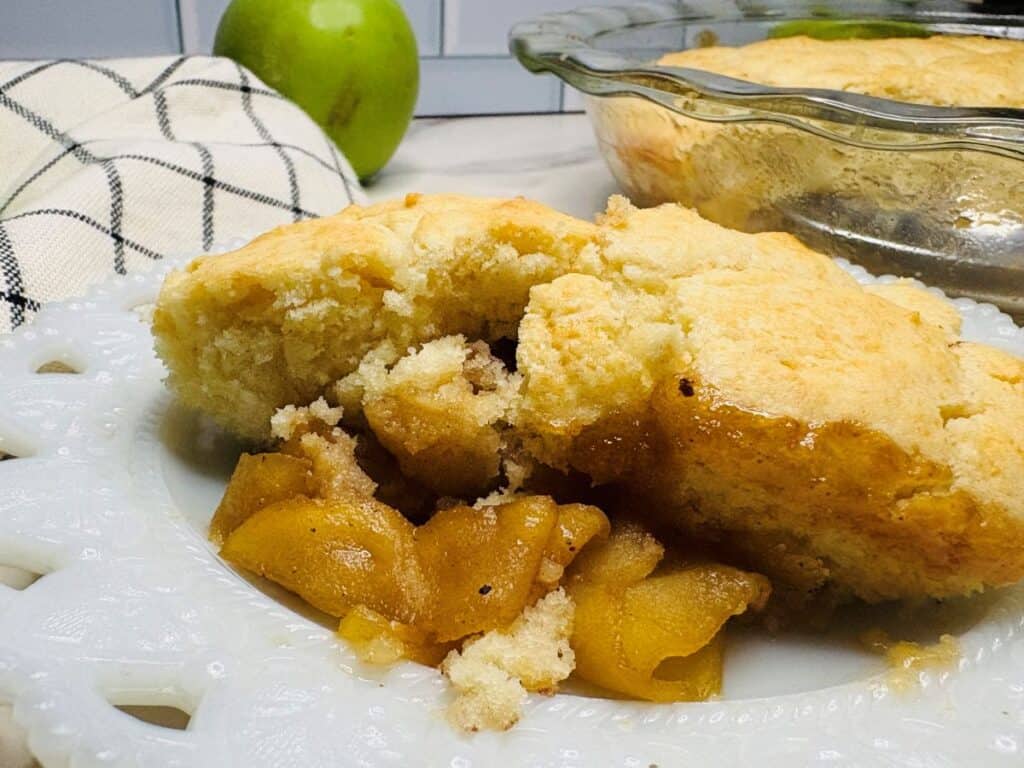A serving of apple cobbler with golden biscuit topping on a white plate, showing baked apples underneath. A glass baking dish, apple, and checkered towel are in the background.