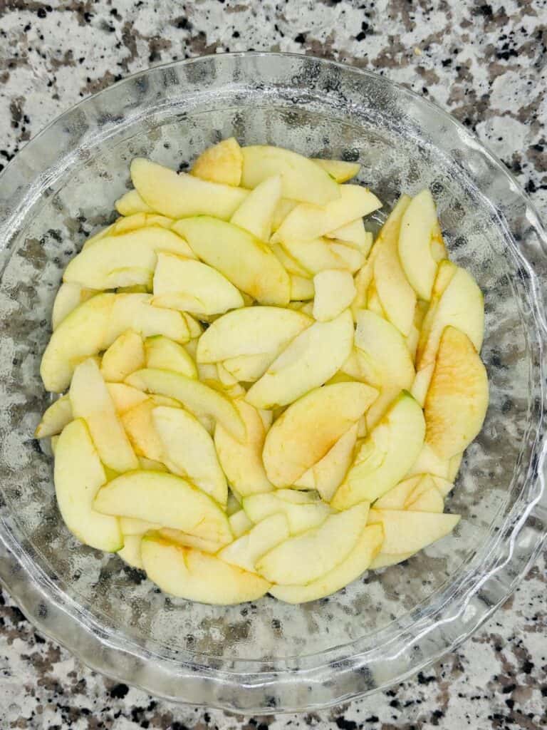 A glass pie dish filled with thinly sliced apples arranged in layers, ready for baking. The dish is placed on a speckled countertop.