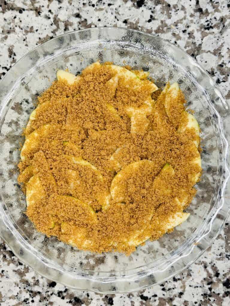 A glass pie dish filled with sliced apples layered and topped with a generous amount of brown sugar, sitting on a speckled countertop.