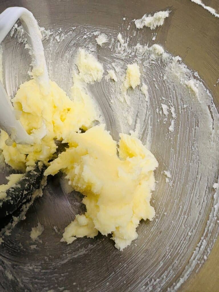Butter and sugar are being creamed together in a metal mixing bowl, with a white mixer paddle partly visible, creating a smooth, pale yellow mixture.