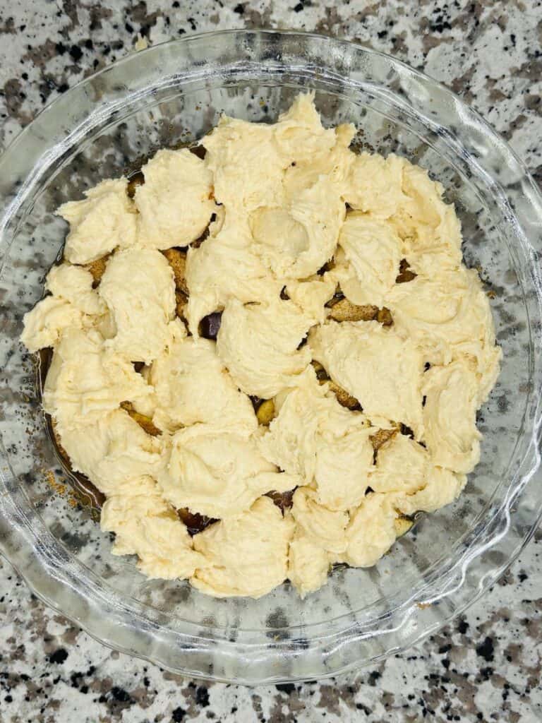 A glass pie dish sits on a granite countertop, filled with unbaked cookie dough dollops covering chunks of what appears to be brownie or cake.