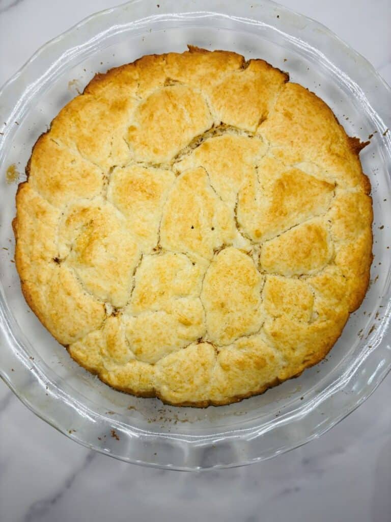 A golden-brown cobbler with a biscuit topping in a clear glass pie dish, set on a white marble surface.