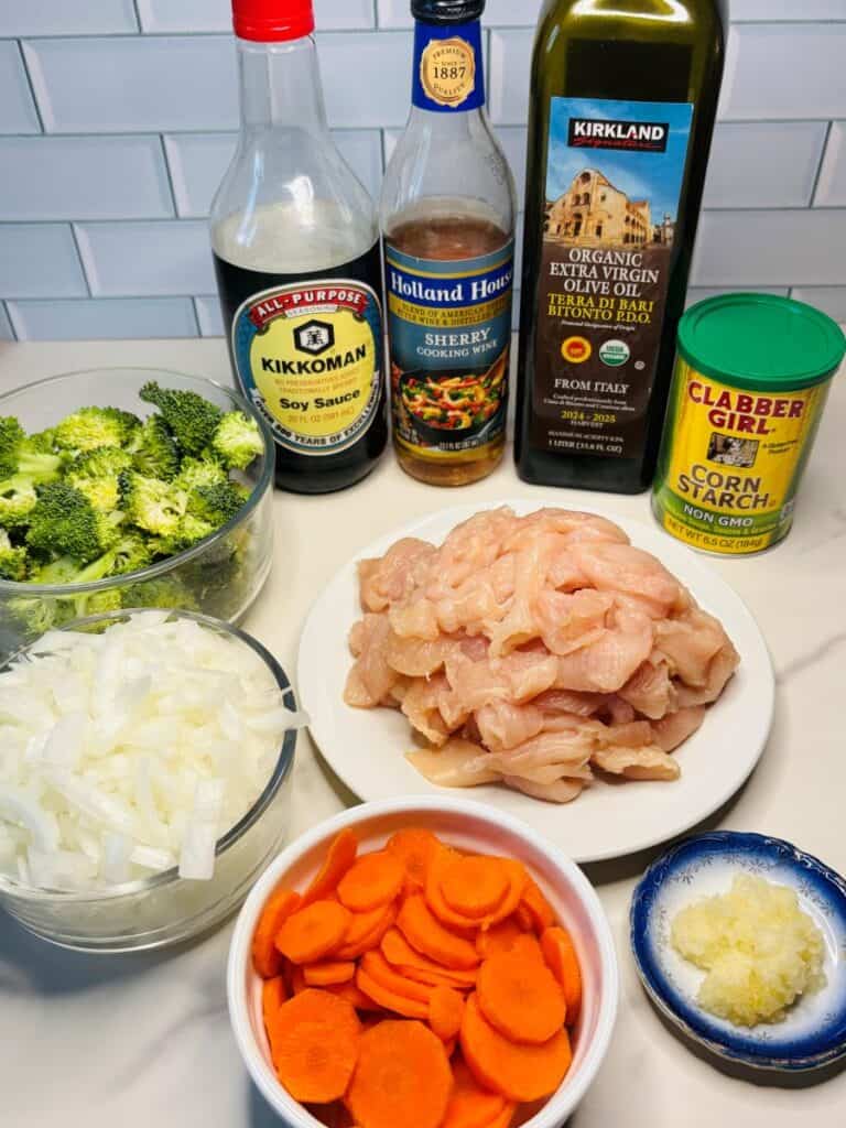 Ingredients for a stir-fry: sliced chicken, broccoli, carrots, chopped onion, soy sauce, sherry cooking wine, olive oil, corn starch, and minced garlic, arranged on a white surface with a white tile background.