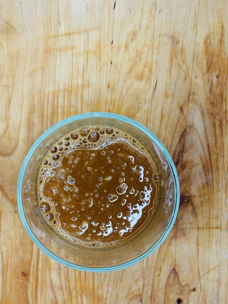 A clear glass bowl filled with a brown, bubbly liquid sits on a light wooden surface with visible grain patterns.