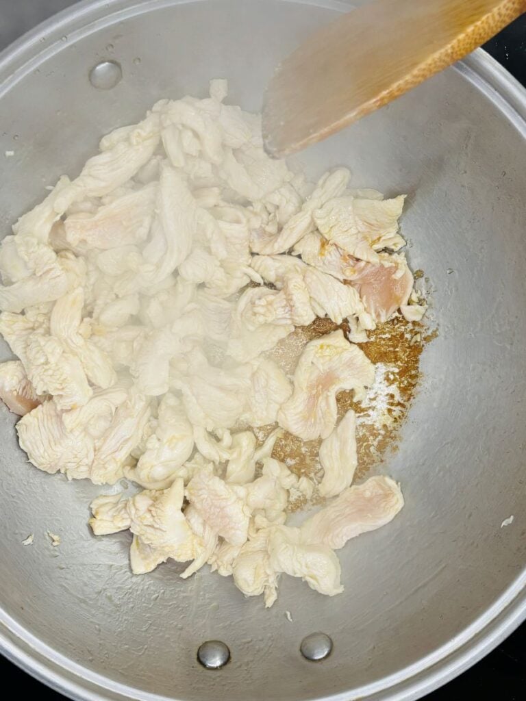 Sliced raw chicken pieces cooking in a large metal pan, with steam rising and a wooden spatula stirring the meat.