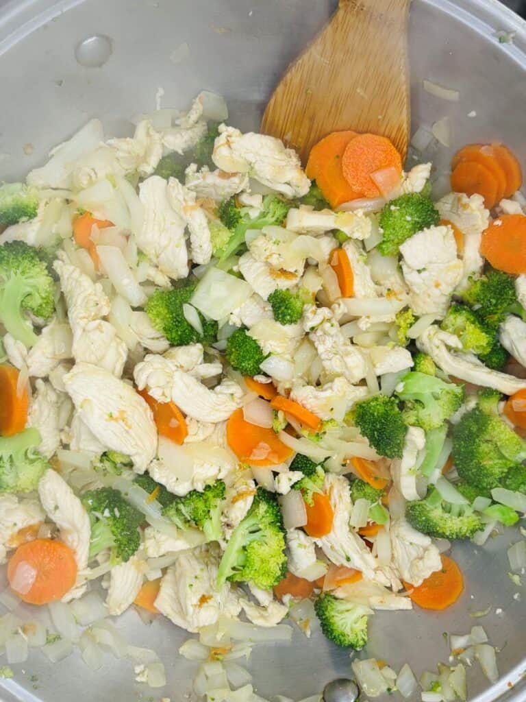 Chicken stir-fry in a pan with broccoli, carrots, onions, and a wooden spatula. The vegetables and chicken are freshly cooked and mixed together. Steam is rising from the hot dish.