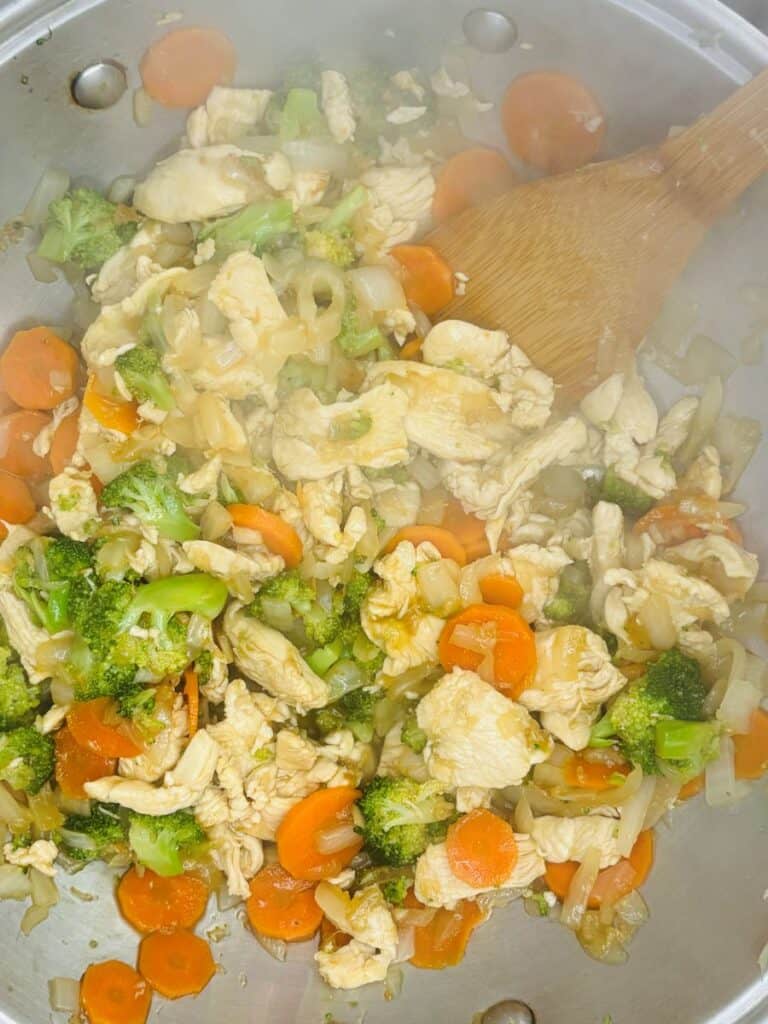 A metal pan filled with stir-fried chicken strips, broccoli, carrots, and onions, being stirred with a wooden spoon. Steam rises from the hot food.