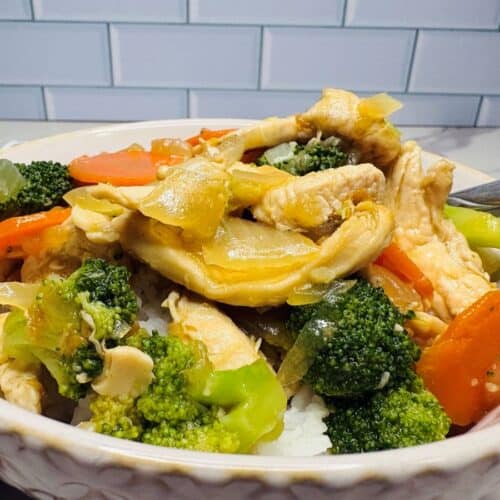 A bowl of chicken stir-fry with broccoli, carrots, and onions served over white rice, placed in front of a white tiled wall.