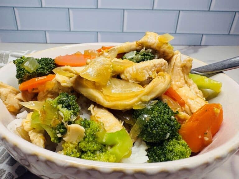 A bowl of chicken stir-fry with broccoli, carrots, and onions served over white rice, placed in front of a white tiled wall.