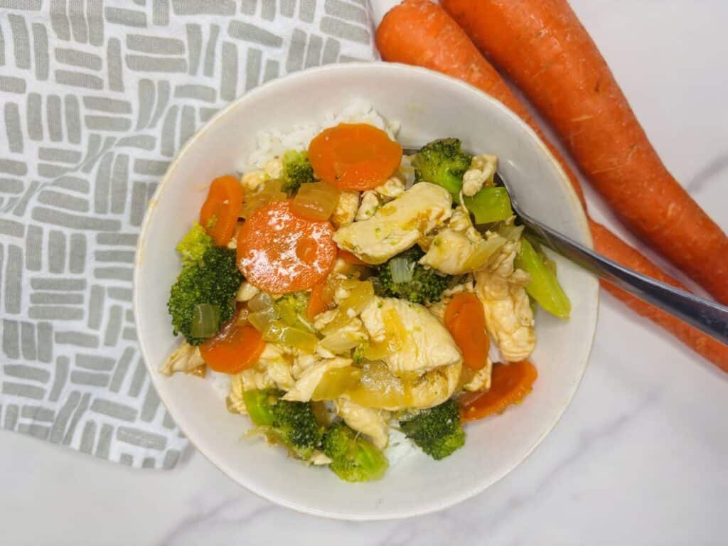 A bowl of chicken stir-fry with broccoli, carrots, and rice on a white surface, next to two whole carrots and a patterned cloth.