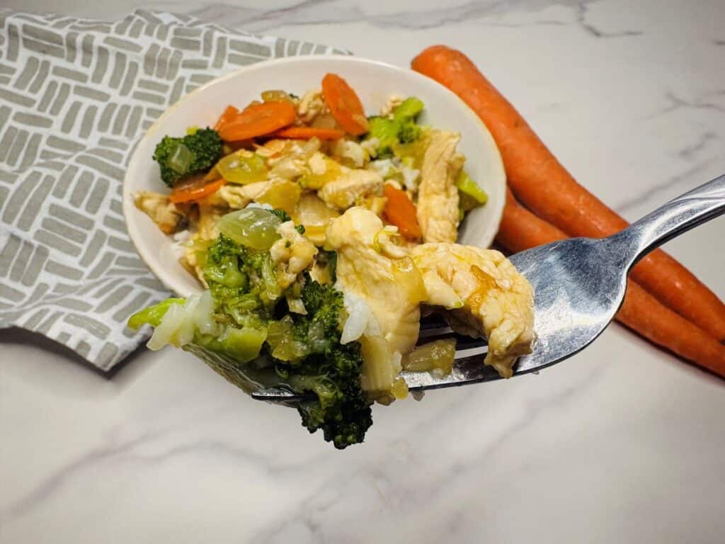 A fork holds a bite of stir-fried chicken, broccoli, and carrots above a bowl of the same dish. Two whole carrots and a patterned napkin are nearby on a marble surface.