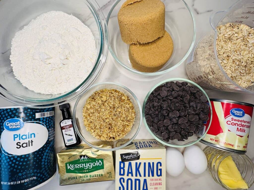 Baking ingredients on a counter: flour, salt, brown sugar, oats, walnuts, chocolate chips, sweetened condensed milk, butter, baking soda, vanilla extract, and two eggs.