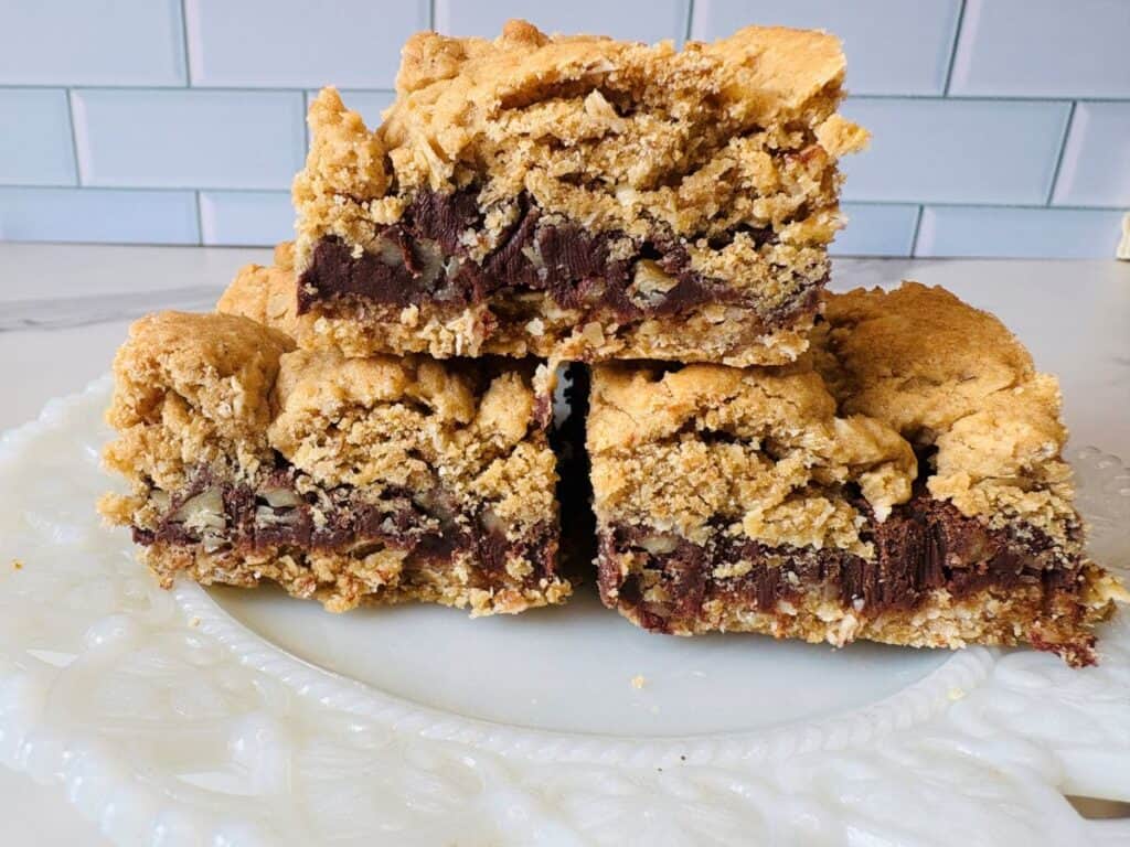 Three thick, crumbly blondie bars with chocolate chunks and nuts are stacked on a white, ornate plate in front of a light blue tiled wall.