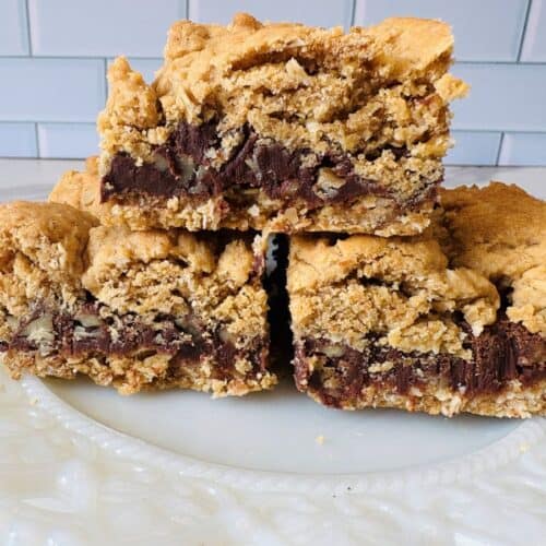 Three thick, crumbly blondie bars with chocolate chunks and nuts are stacked on a white, ornate plate in front of a light blue tiled wall.