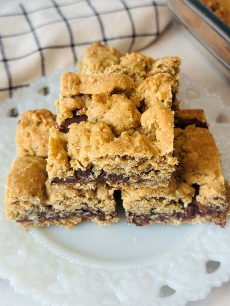 Four oat and chocolate chip cookie bars are stacked on a white decorative plate, with a checkered towel and a glass baking dish in the background.
